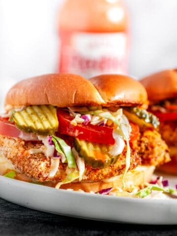 Panko Crusted Spicy Chicken Sandwiches with coleslaw, tomato, and pickles on white plate with hot sauce bottle in background