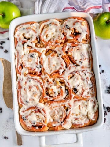 Apple Currant Cinnamon Rolls in white baking dish with gold pie server on white marble