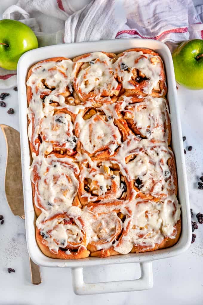 Apple Currant Cinnamon Rolls in white baking dish with gold pie server on white marble