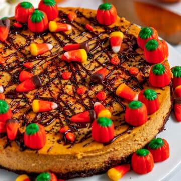 Halloween Pumpkin Cheesecake topped with chocolate drizzle and candy corn on white cake plate with gold pie server