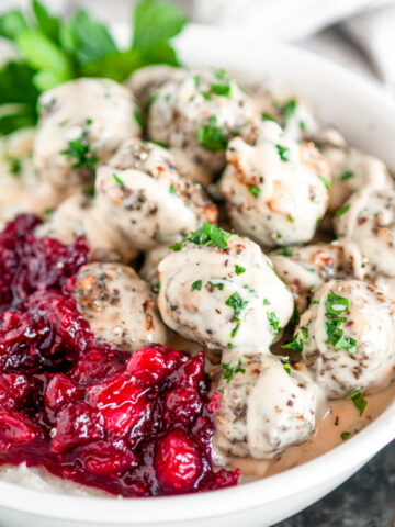 Easy Baked Swedish Meatballs in white bowl with mashed potatoes, cranberry sauces and parsley on white marble