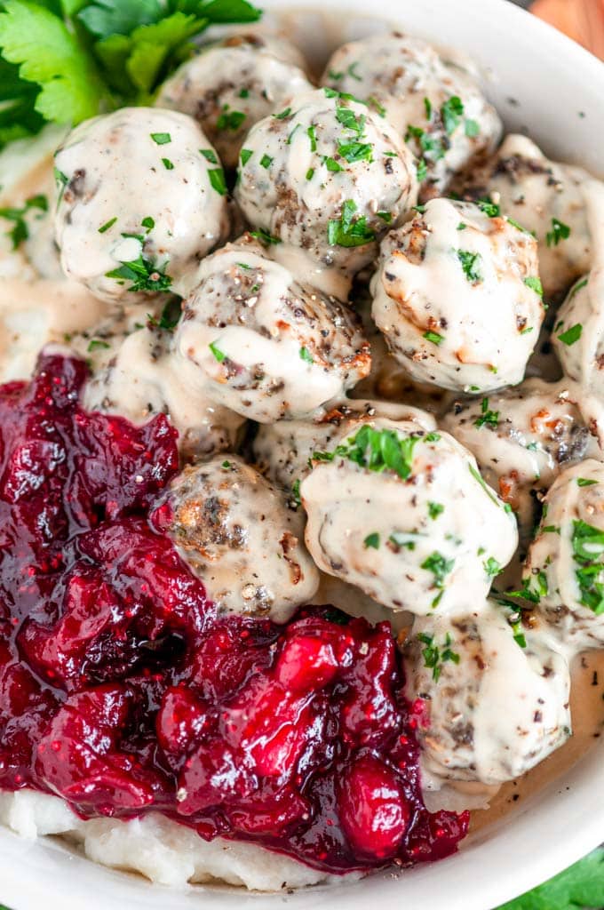 Easy Baked Swedish Meatballs in white bowl with mashed potatoes, cranberry sauces and parsley on white marble