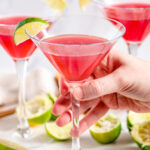 Classic Cosmopolitan Cocktail in martini glasses with lime wedge held in hand on white marble