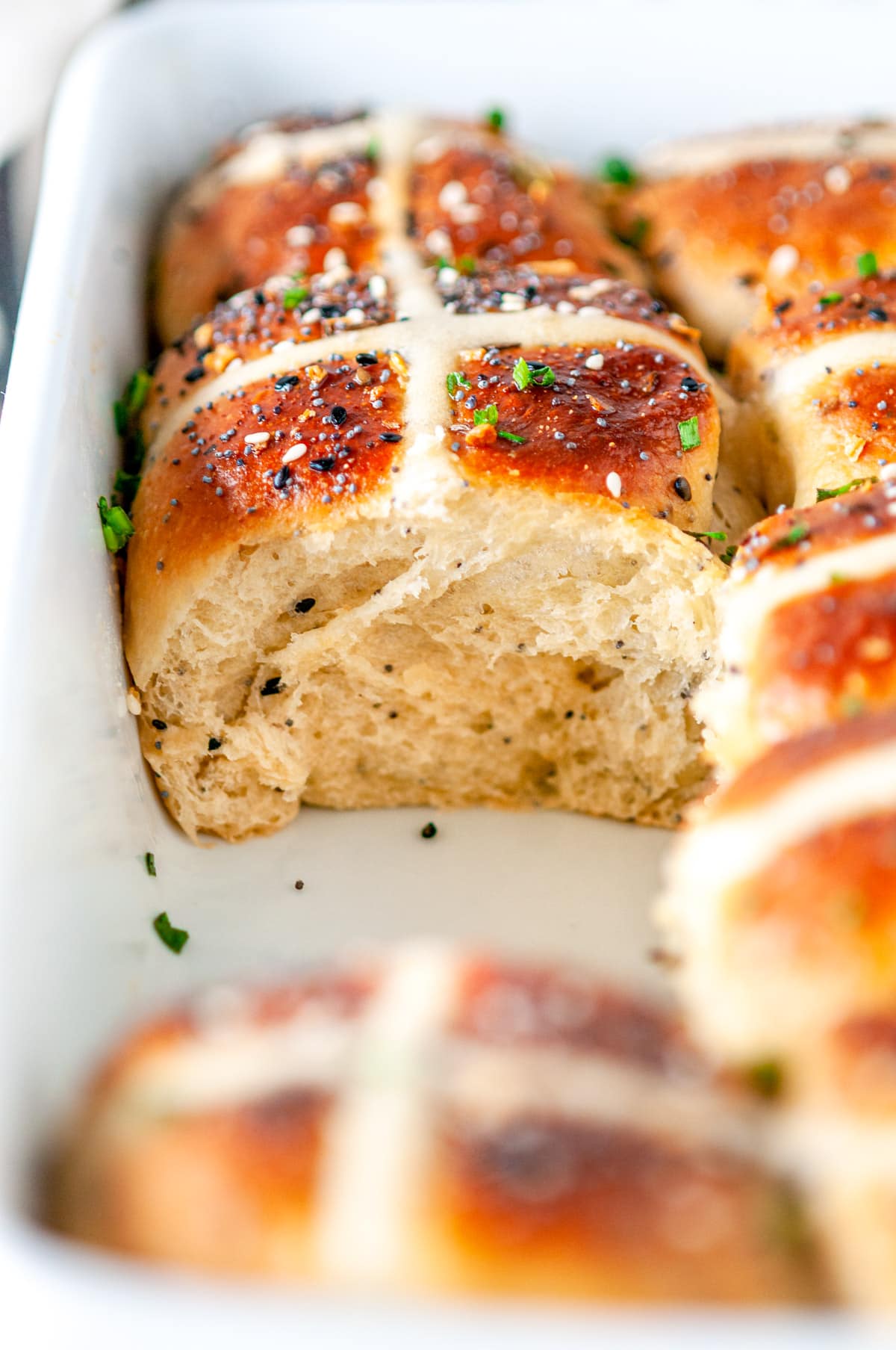 Everything Bagel Hot Cross Buns in white baking dish with sliced chives, one bun removed, on white marble side view close up