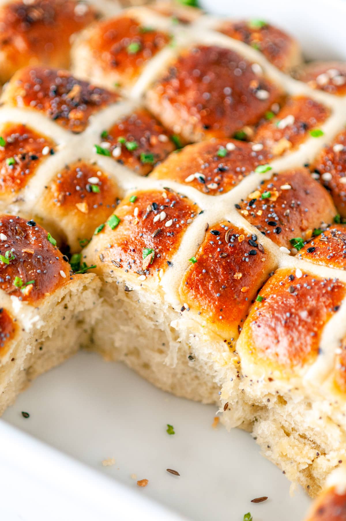 Everything Bagel Hot Cross Buns in white baking dish with sliced chives, one bun removed, on white marble side view close up