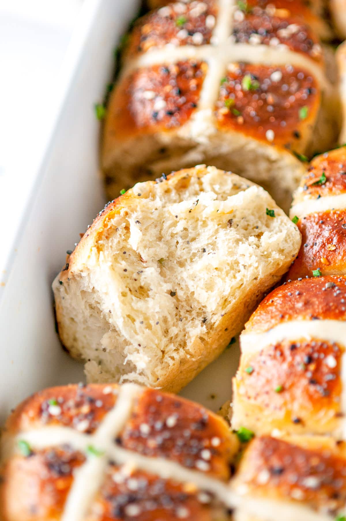 Everything Bagel Hot Cross Buns in white baking dish with sliced chives, one bun on it's side, on white marble close up