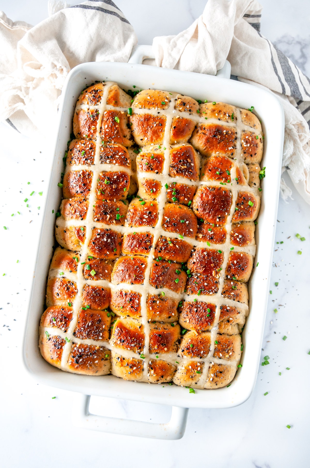 Everything Bagel Hot Cross Buns in white baking dish with sliced chives on white marble overhead view