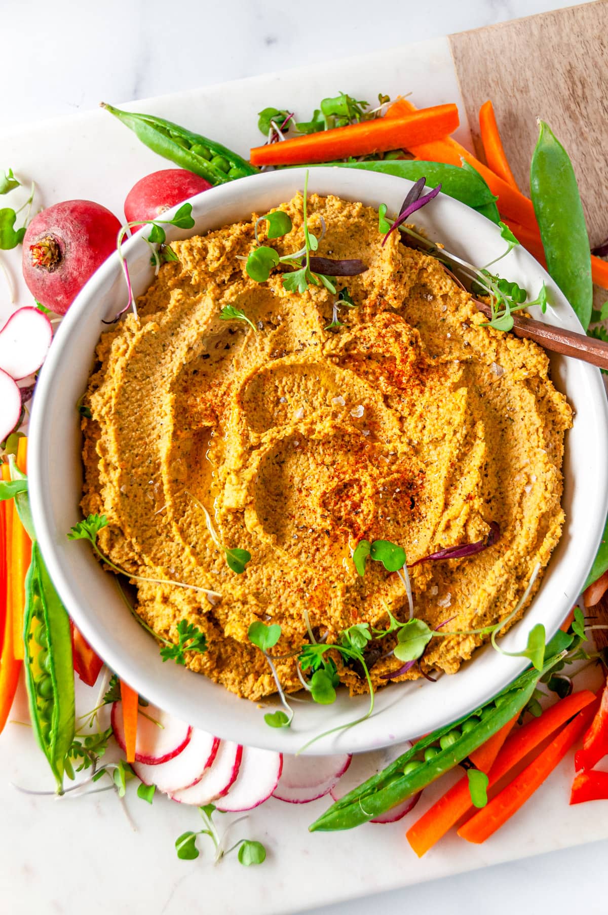 Easy Roasted Vegetable Hummus in white bowl topped with microgreens and surrounded by fresh veggies on white marble overhead view