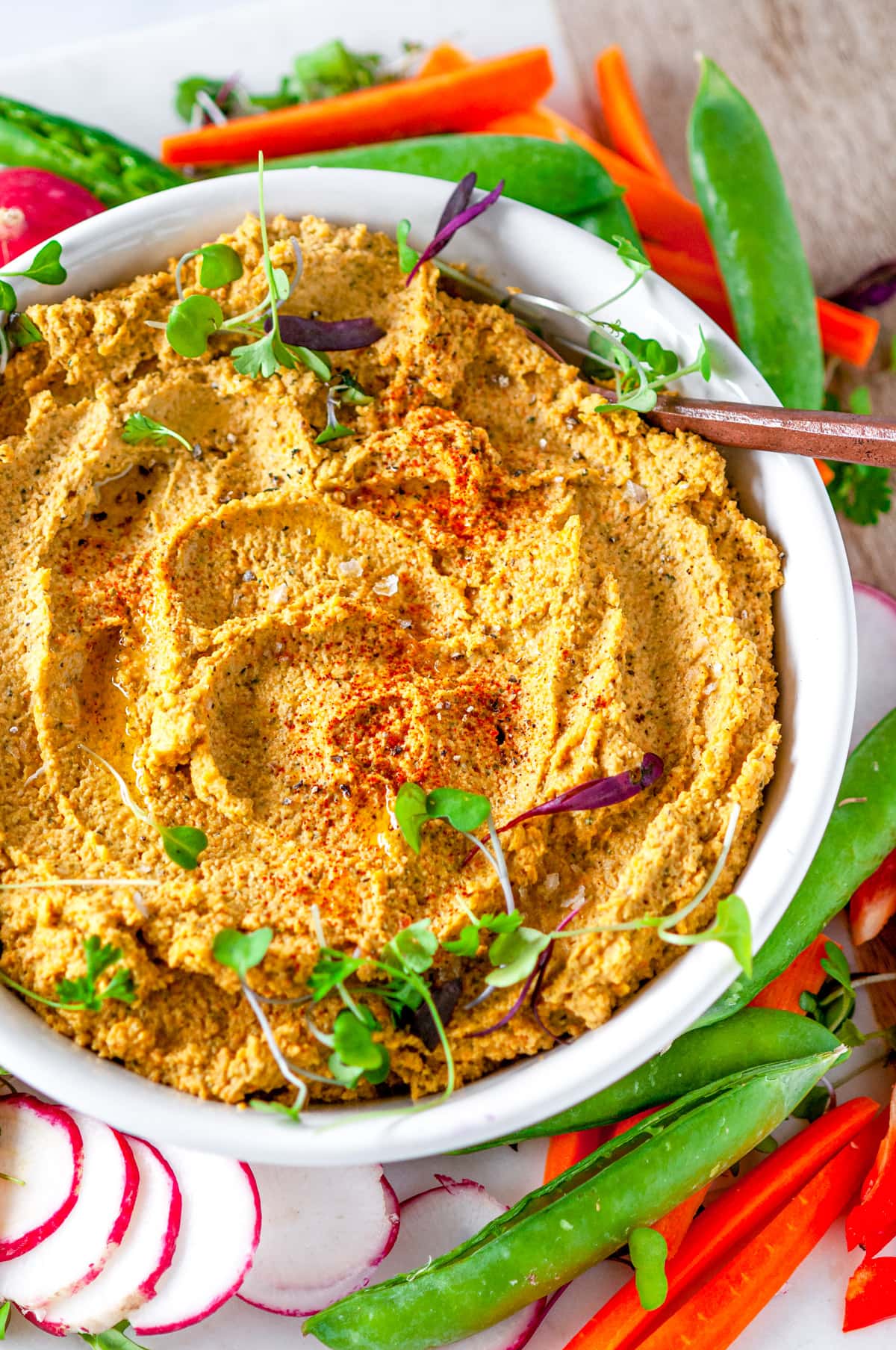 Easy Roasted Vegetable Hummus in white bowl topped with microgreens and surrounded by fresh veggies on white marble close up overhead view