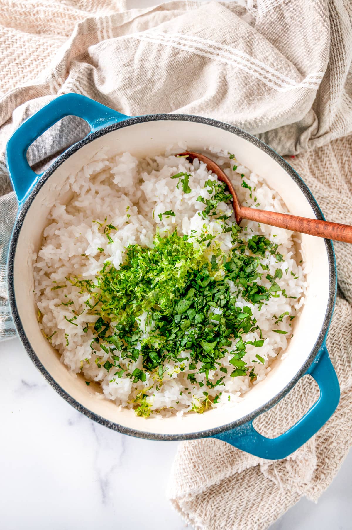 Cilantro lime rice in medium blue stock pot with wooden serving spoon before mixing together on white marble overhead view