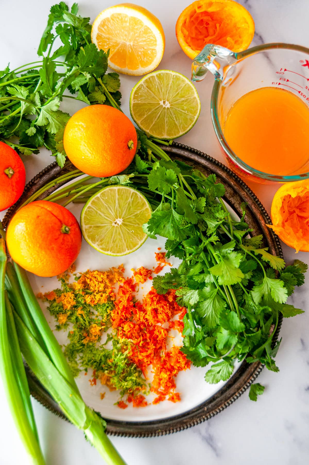 Ingredients for a citrus marinade on white marble: juiced oranges, halved lemons, halved limes, green onion stalks, and cilantro bunch with orange juice and citrus zest.