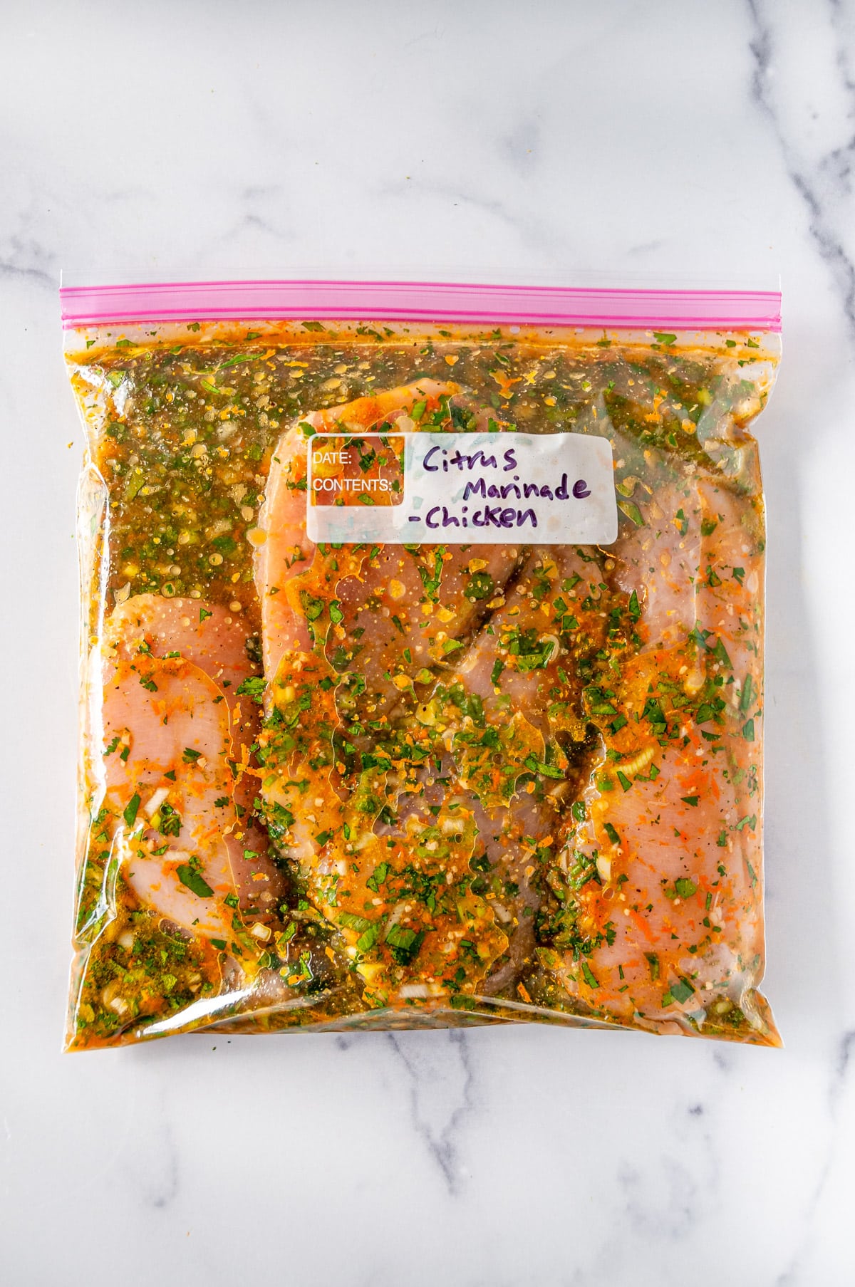 Four raw chicken breasts in a ziploc bag coated in a citrus marinade on white marble