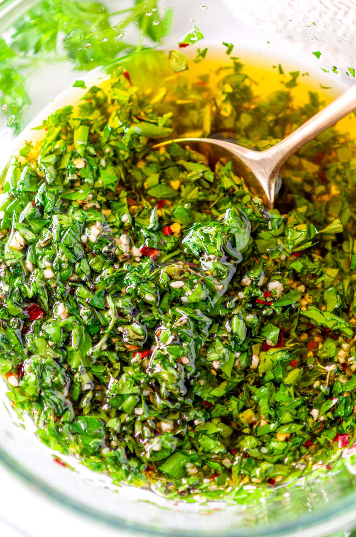 Five Minute Chimichurri Sauce in clear mixing bowl with fresh parsley and gold spoon on white marble