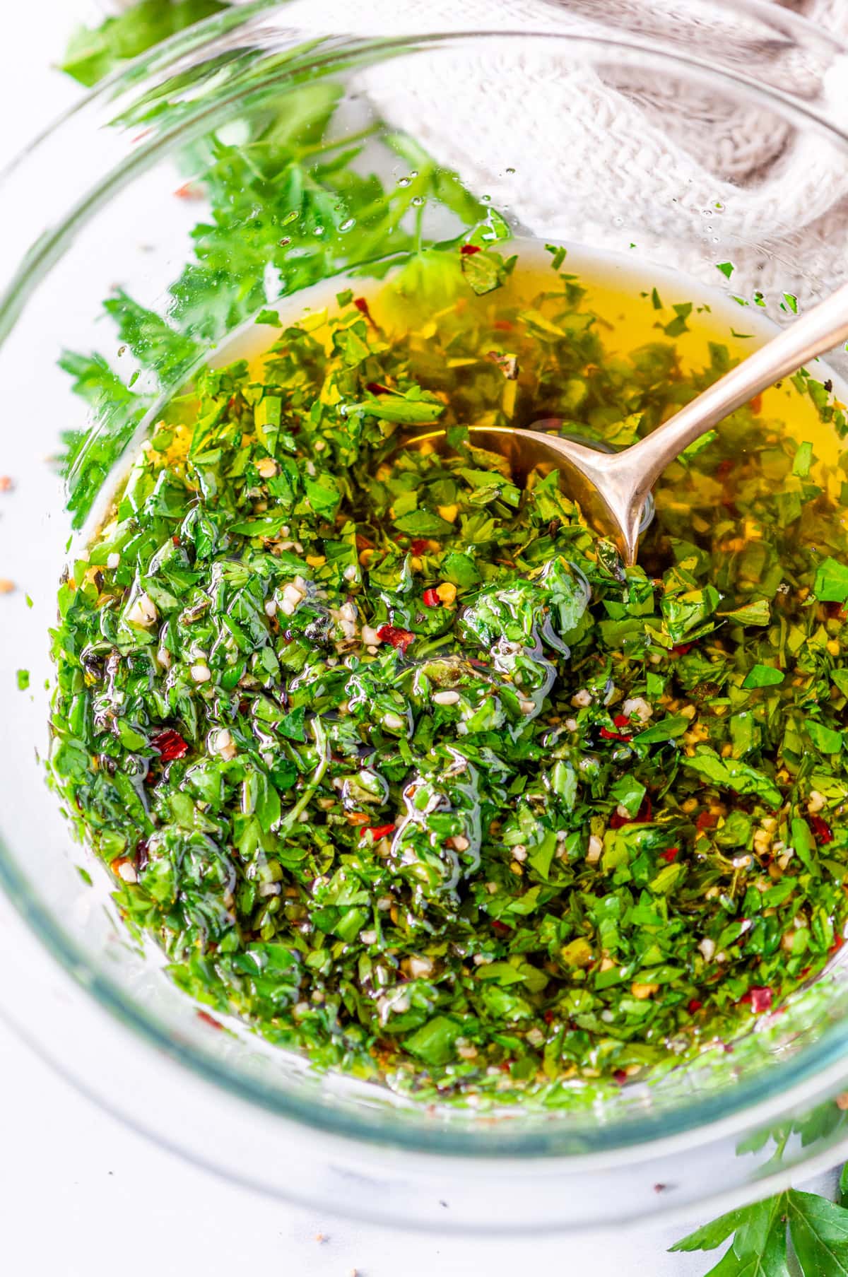 Five Minute Chimichurri Sauce in clear mixing bowl with fresh parsley and gold spoon on white marble
