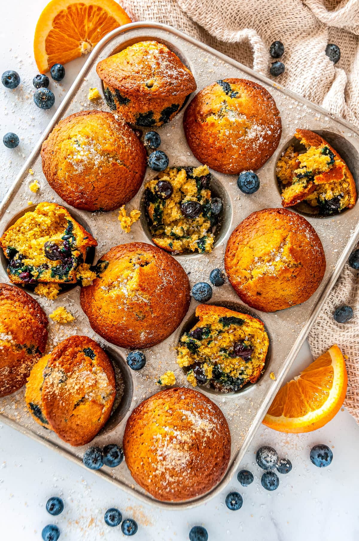 Zesty Orange Blueberry Muffins in silver muffin tray with sliced oranges and fresh blueberries on white marble overhead view