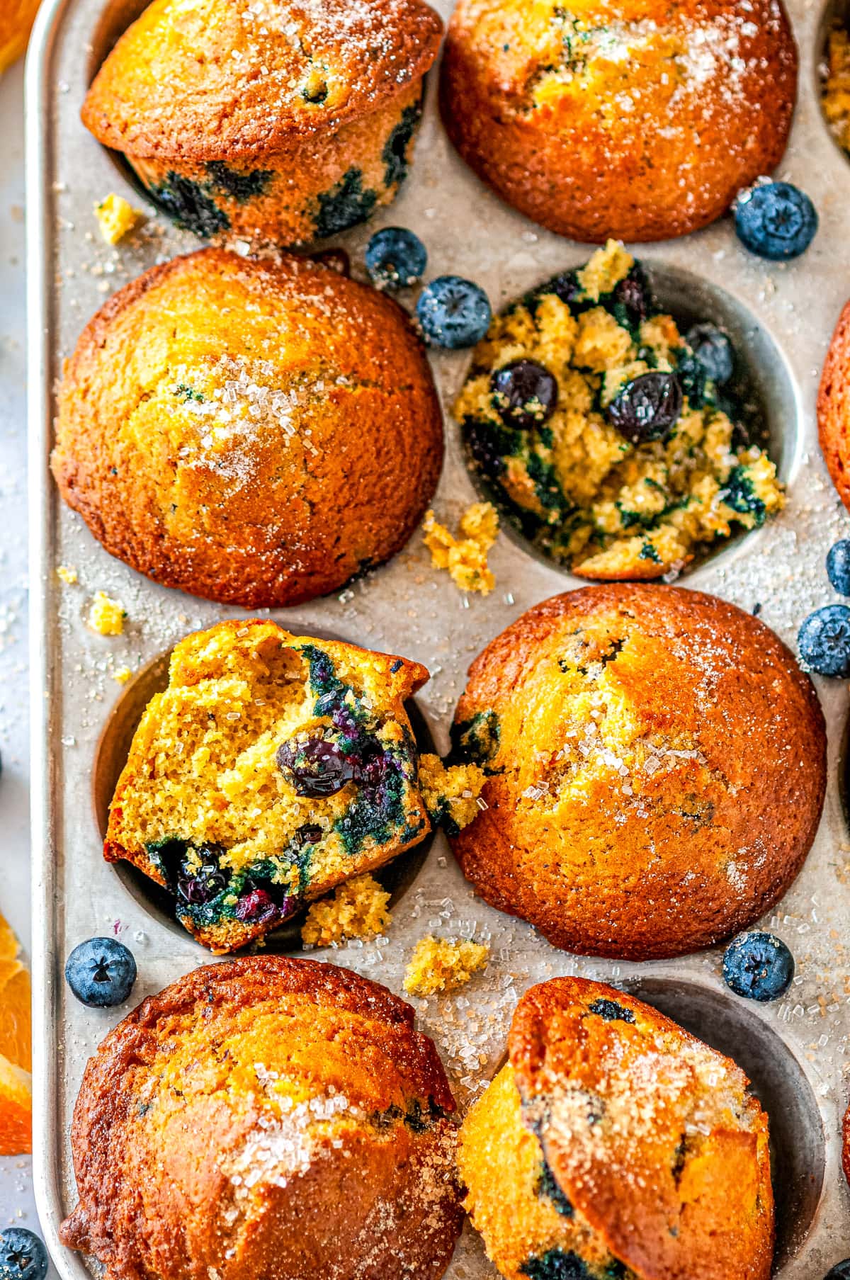 Zesty Orange Blueberry Muffins in silver muffin tray with sliced oranges and fresh blueberries on white marble overhead view close up