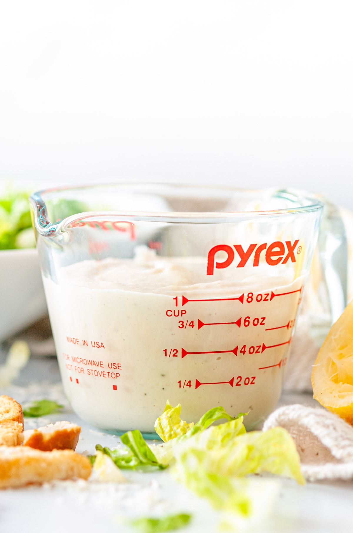 Vegetarian Caesar Salad Dressing in pyrex measuring cup with romaine lettuce, squeezed lemons, and croutons