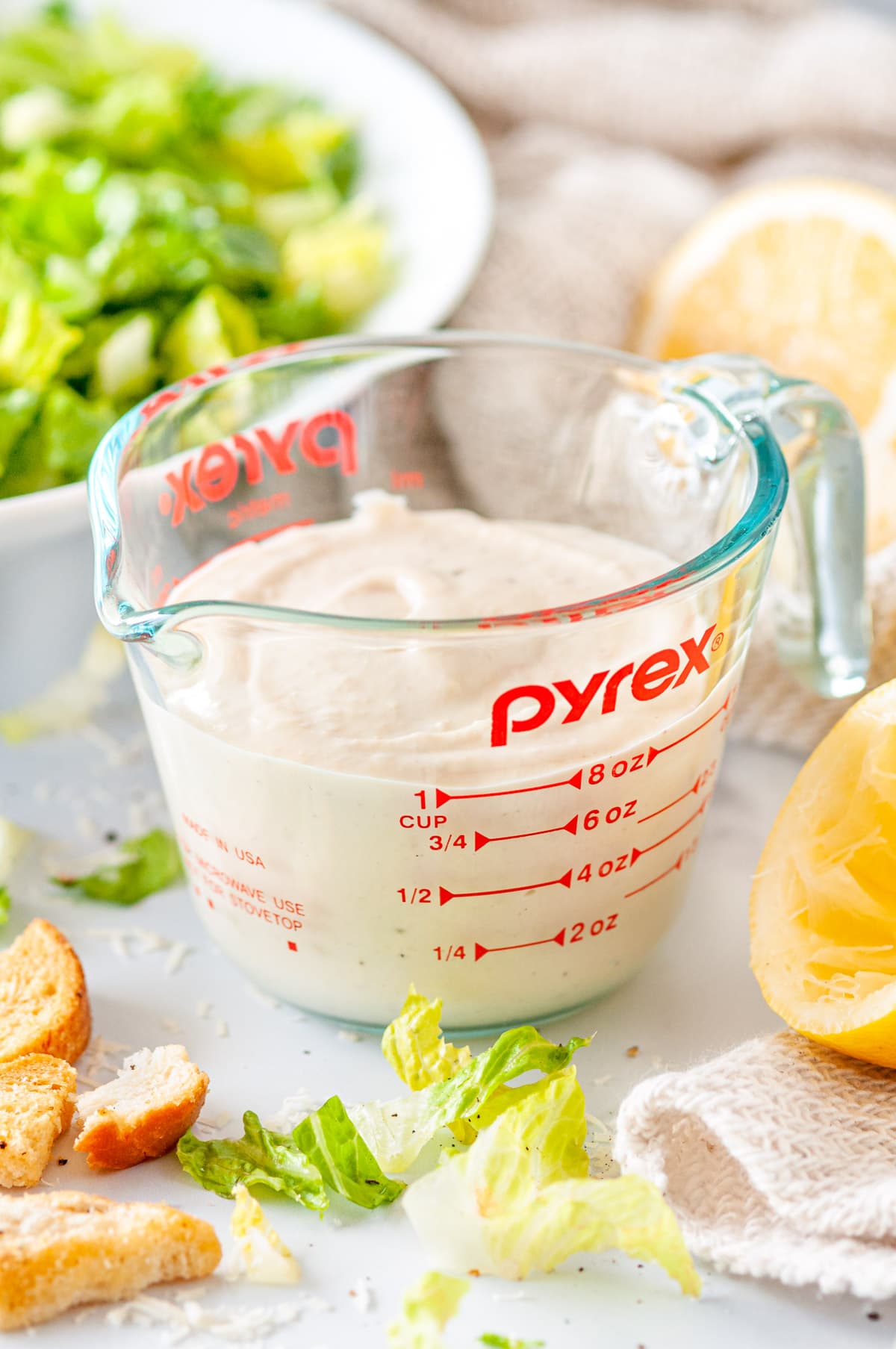 Vegetarian Caesar Salad Dressing in pyrex measuring cup with romaine lettuce, squeezed lemons, and croutons