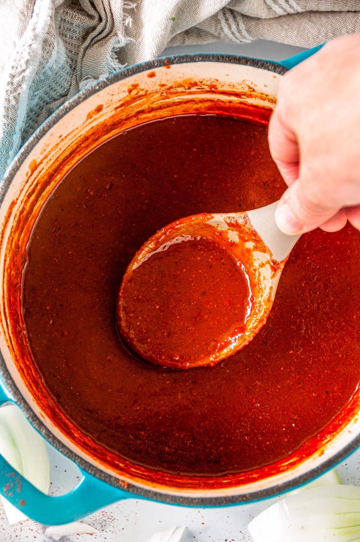 Pantry Friendly Enchilada Sauce in blue enameled saucepan with hand holding gray spoon