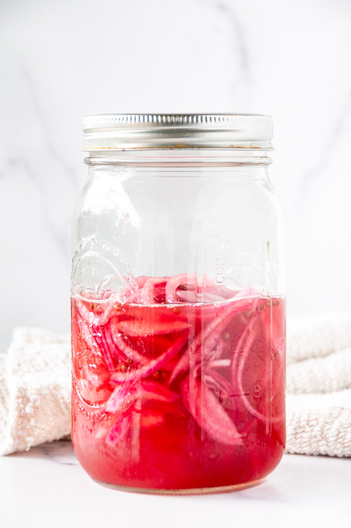 Pickled red onions in a large, wide mouthed mason jar with tea towel on white marble