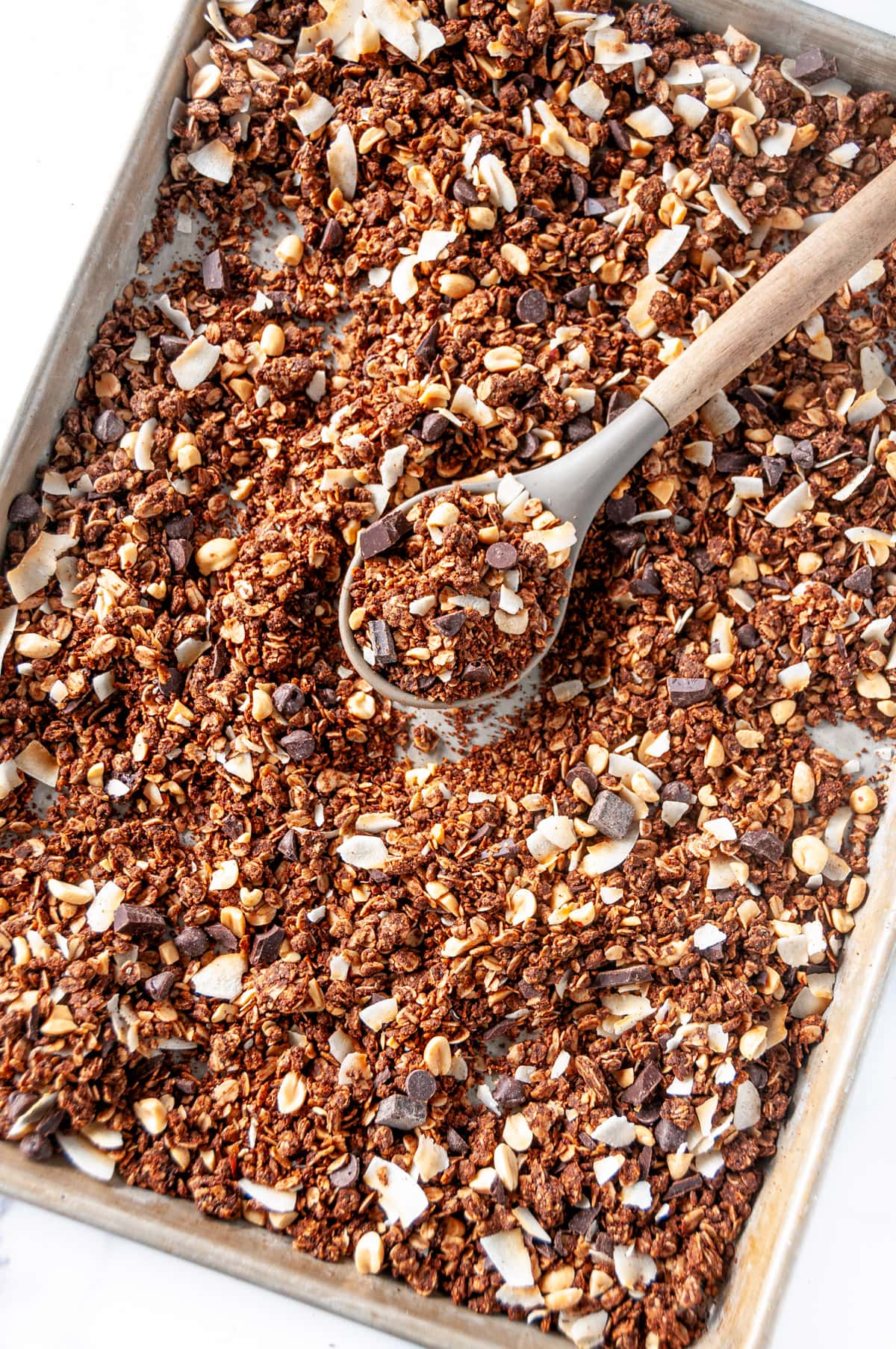 Chocolate peanut butter granola baked on sheet pan with gray silicone and wooden spoon overhead view