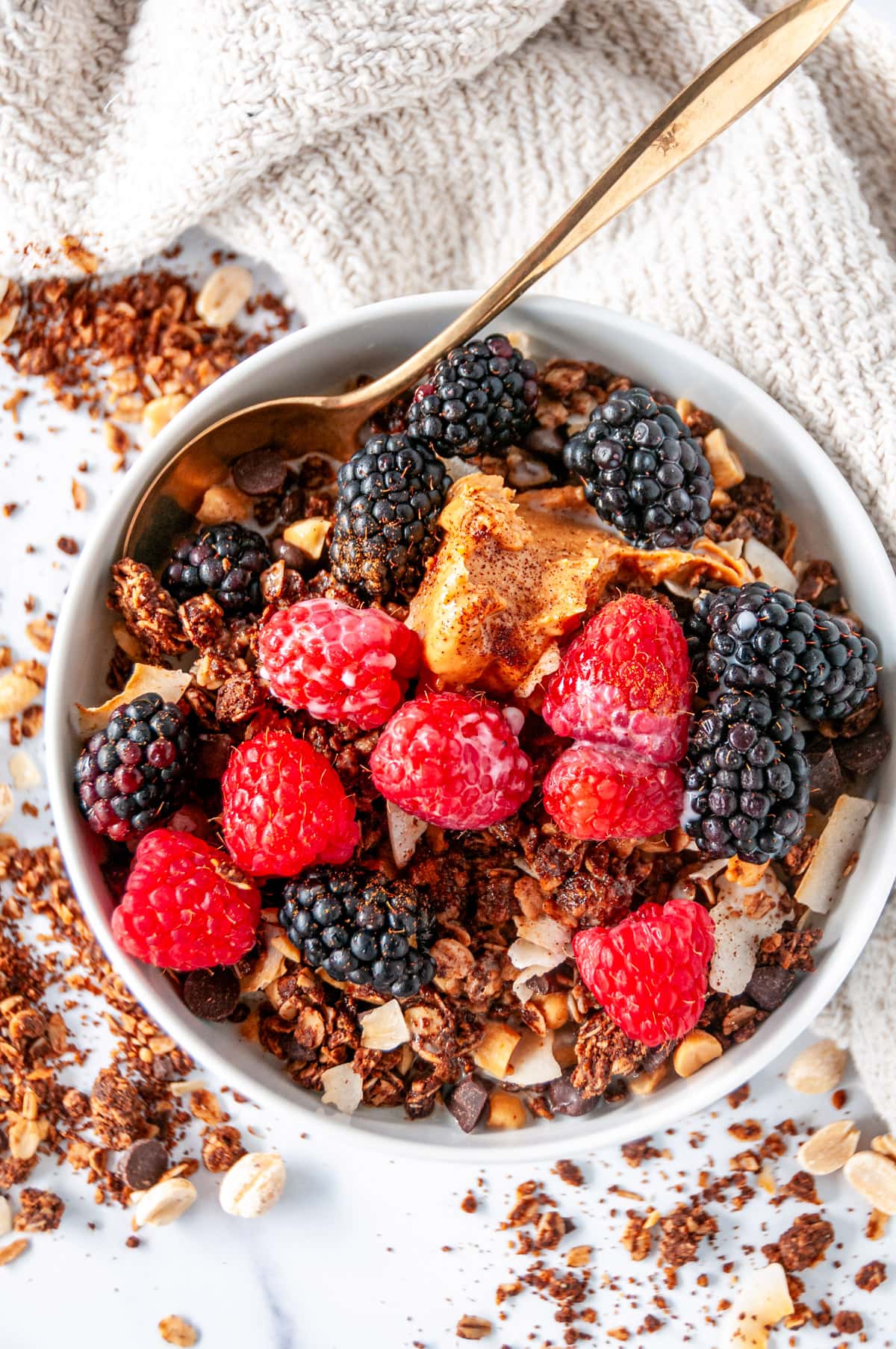 Berries and a scoop of peanut butter on top of chocolate peanut butter granola in a white bowl with a gold spoon on white marble overhead view