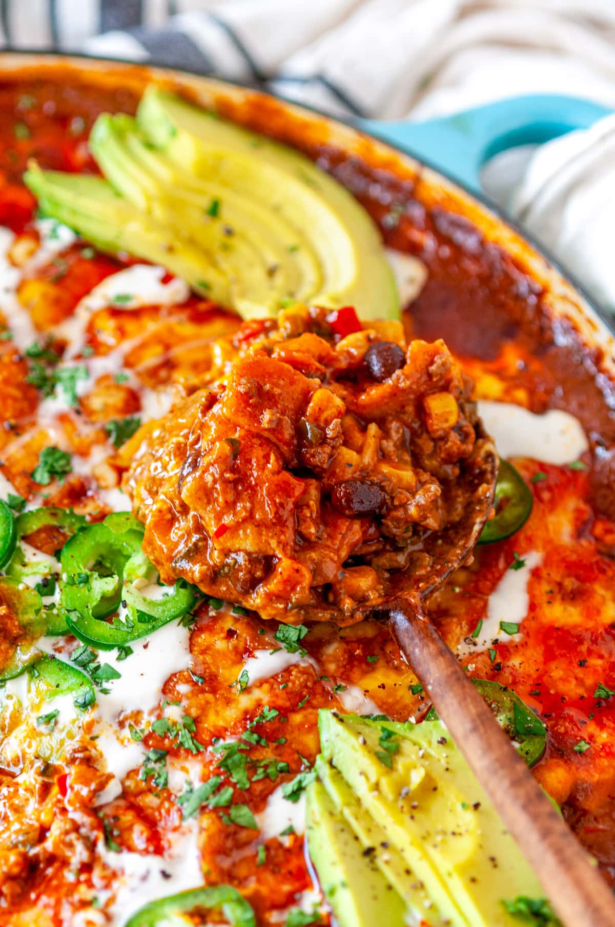 One Pot Red Enchilada Bake with scoop in wooden serving spoon topped with sliced avocado, jalapeno, cilantro, and crema Mexicana