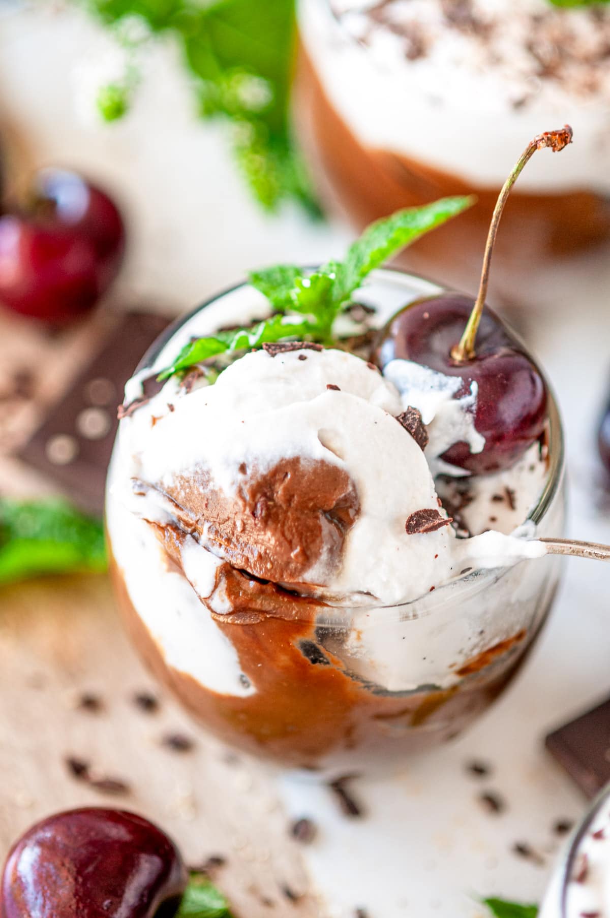 Spoonful of Avocado Chocolate Mousse in small dessert glasses with chocolate, mint, and cherries on white marble close up