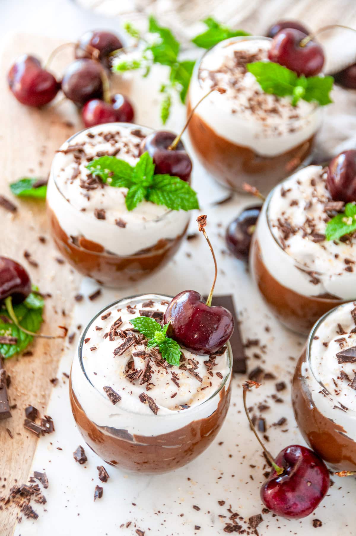 Avocado Chocolate Mousse in small dessert glasses with chocolate, mint, and cherries on white marble