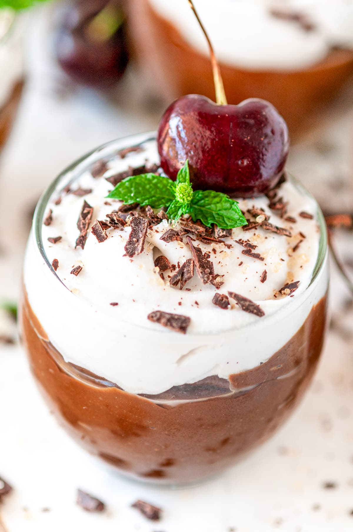 Avocado Chocolate Mousse in small dessert glasses with chocolate, mint, and cherries on white marble close up