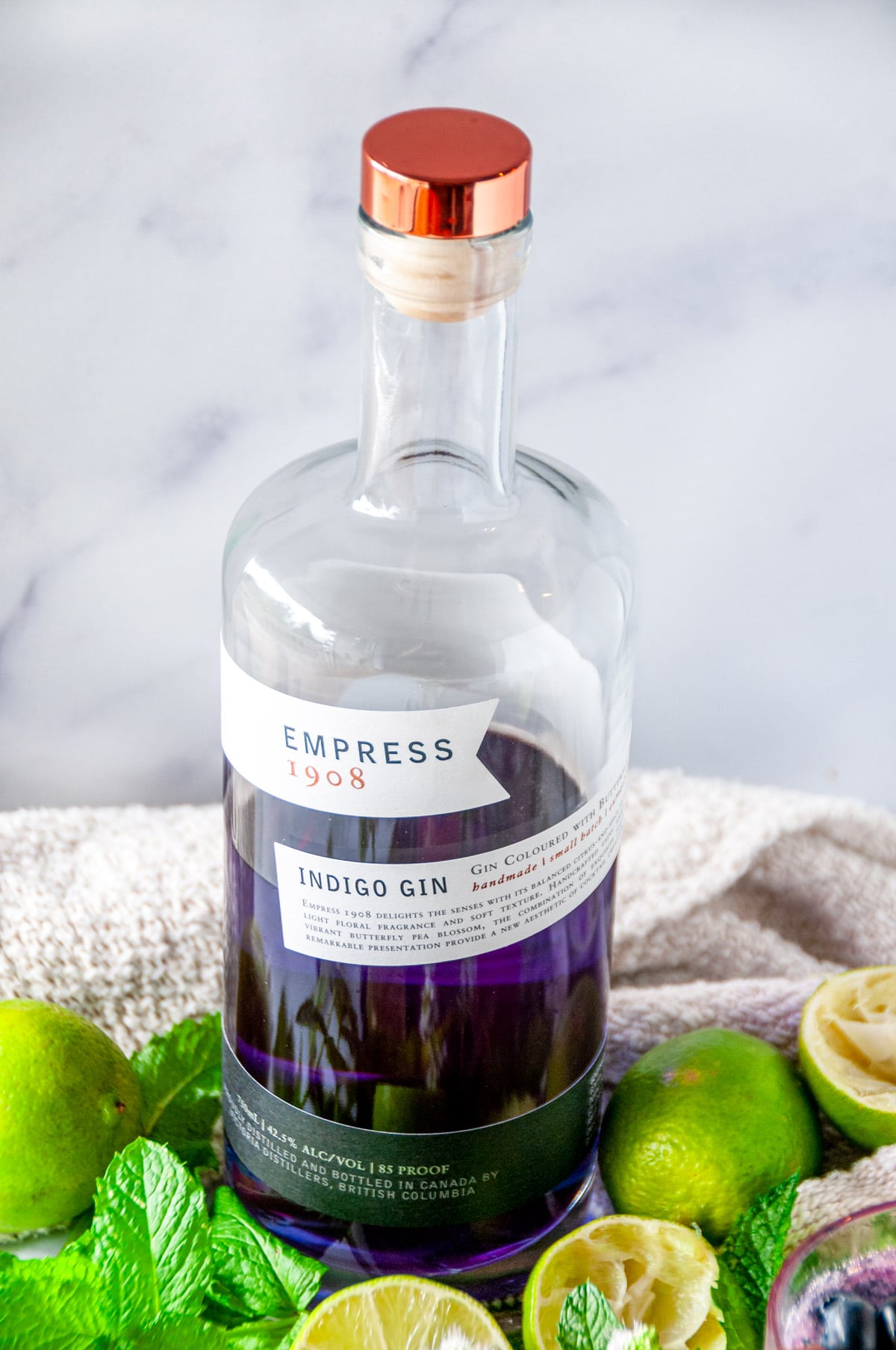 Purple empress gin bottle with white marble background surrounded by mint leaves and lime wedges.