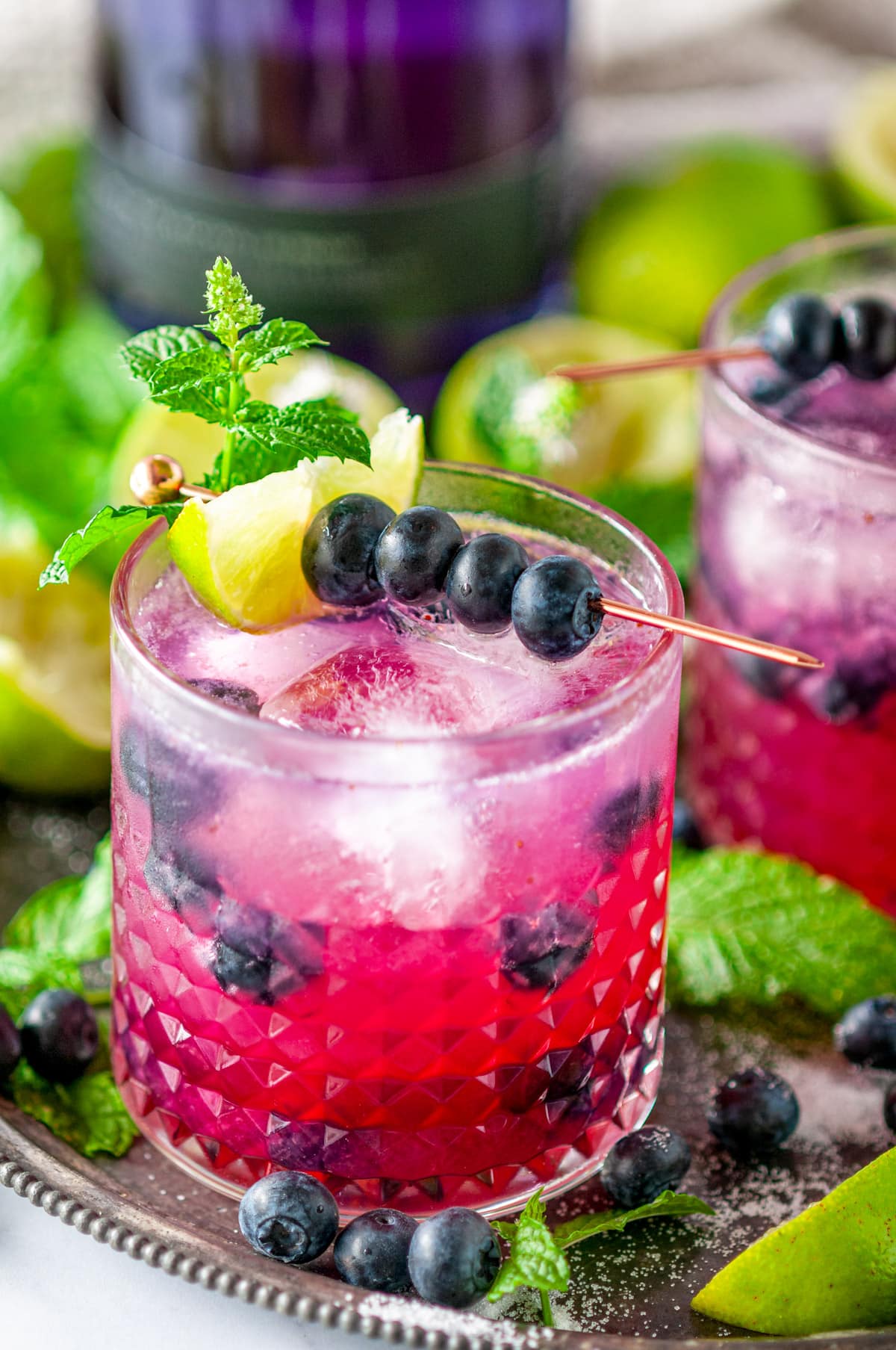 Blueberry GIn Mojito Cocktail on metal plate with fresh mint leaves and lime wedges topped with rose gold fruit garnish