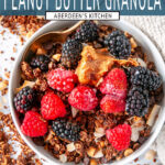 Berries and a scoop of peanut butter on top of chocolate peanut butter granola in a white bowl with a gold spoon on white marble overhead view - teal rectangle overlay with white text
