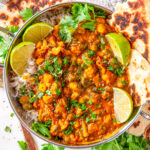 Lentil Chickpea Veggie Curry in metal bowl with limes, cilantro and fresh naan bread on white marble