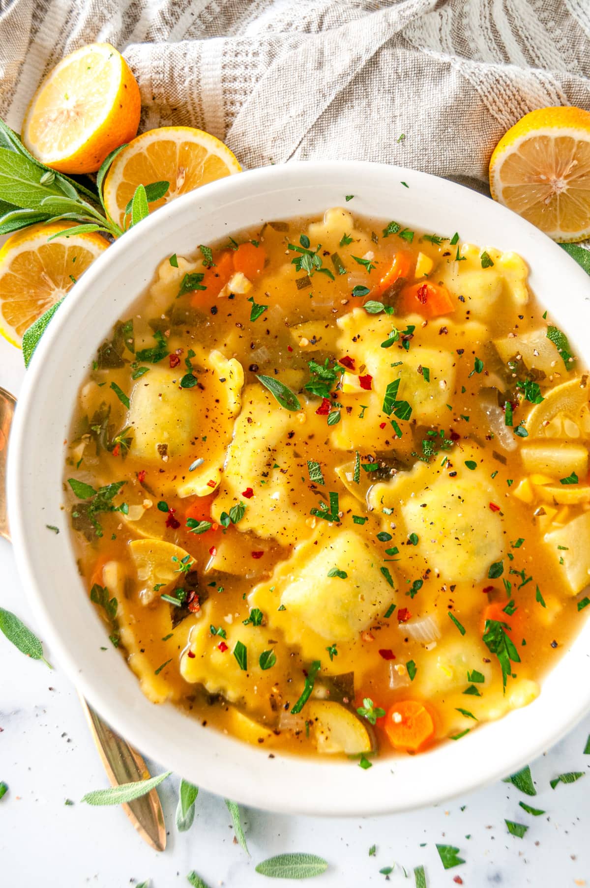 One Pot Ravioli Soup in white bowl with lemons and sage on white marble