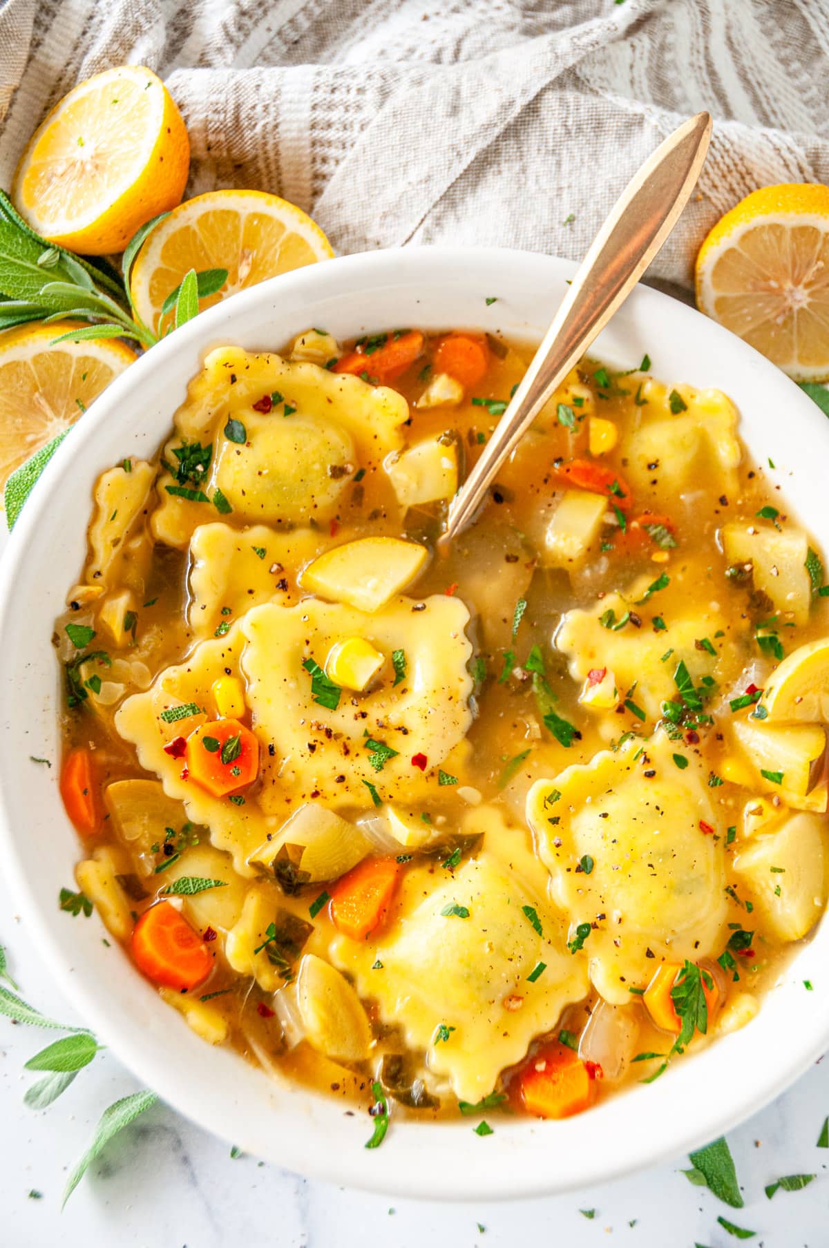 One Pot Ravioli Soup in white bowl with gold spoon, lemons, and sage on white marble