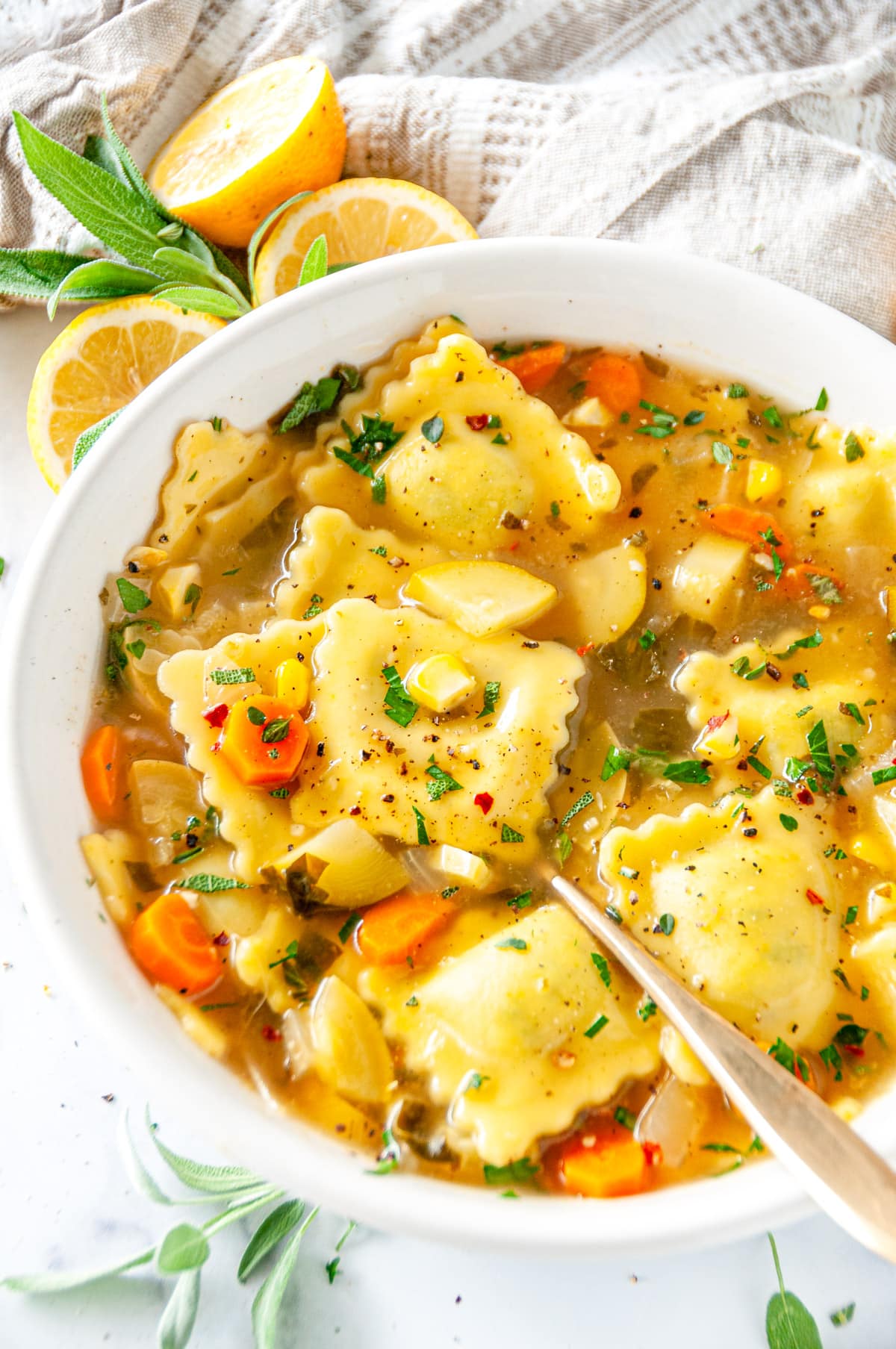One Pot Ravioli Soup in white bowl with gold spoon, lemons, and sage on white marble