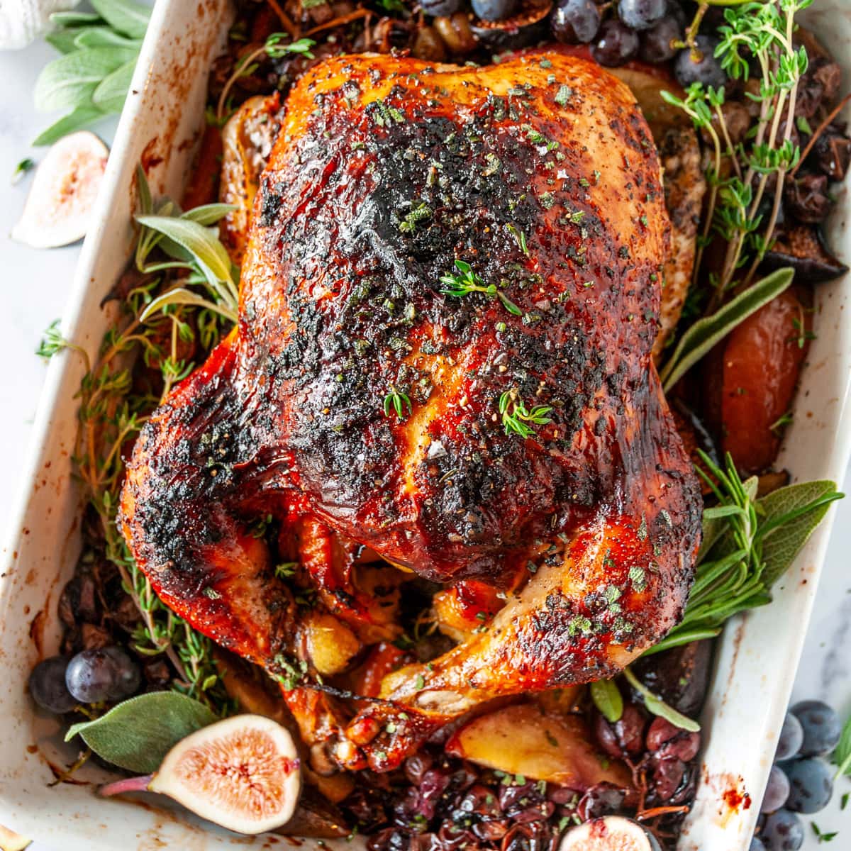 Autumn Apple Cider Roasted Chicken in white baking dish with figs, grapes, and herbs on white marble