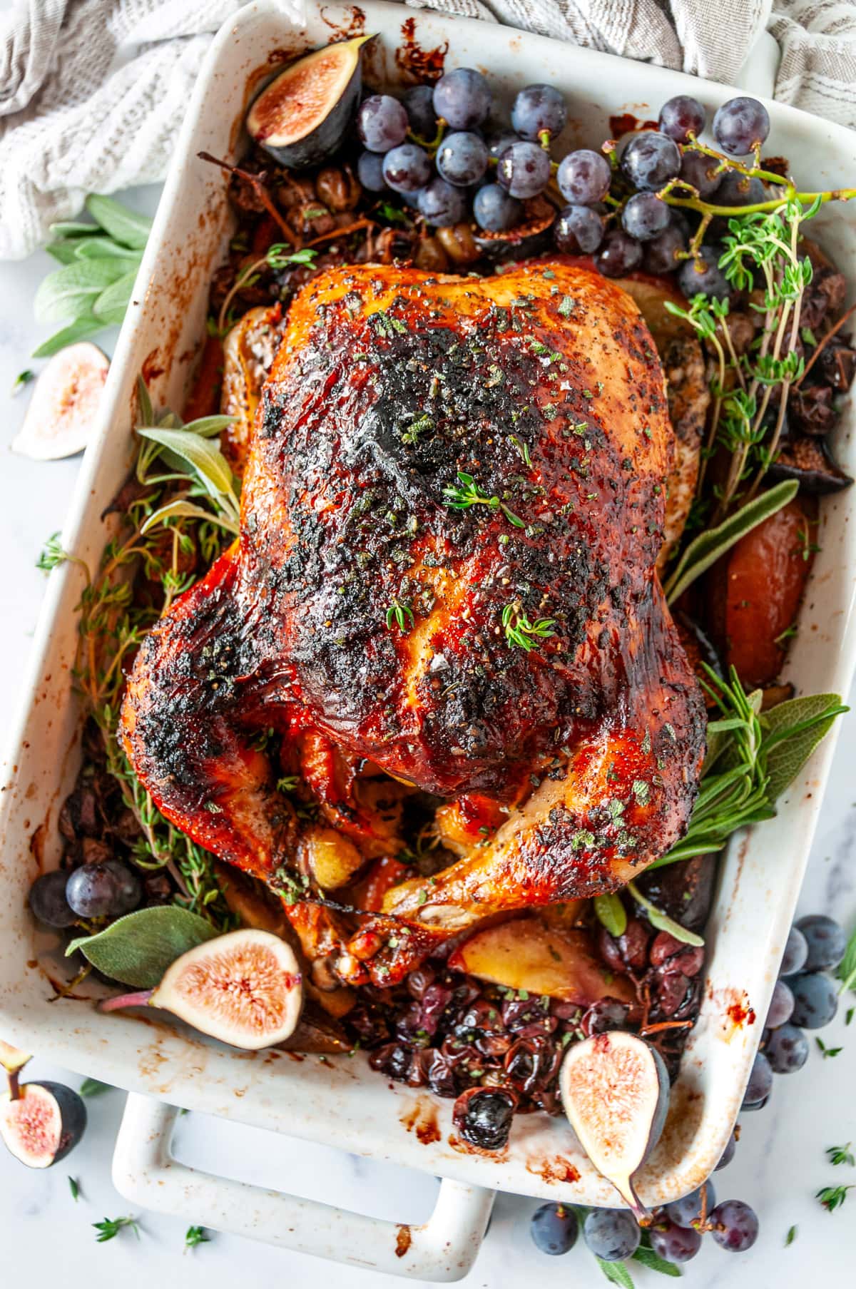 Autumn Apple Cider Roasted Chicken in white baking dish with figs, grapes, and herbs on white marble