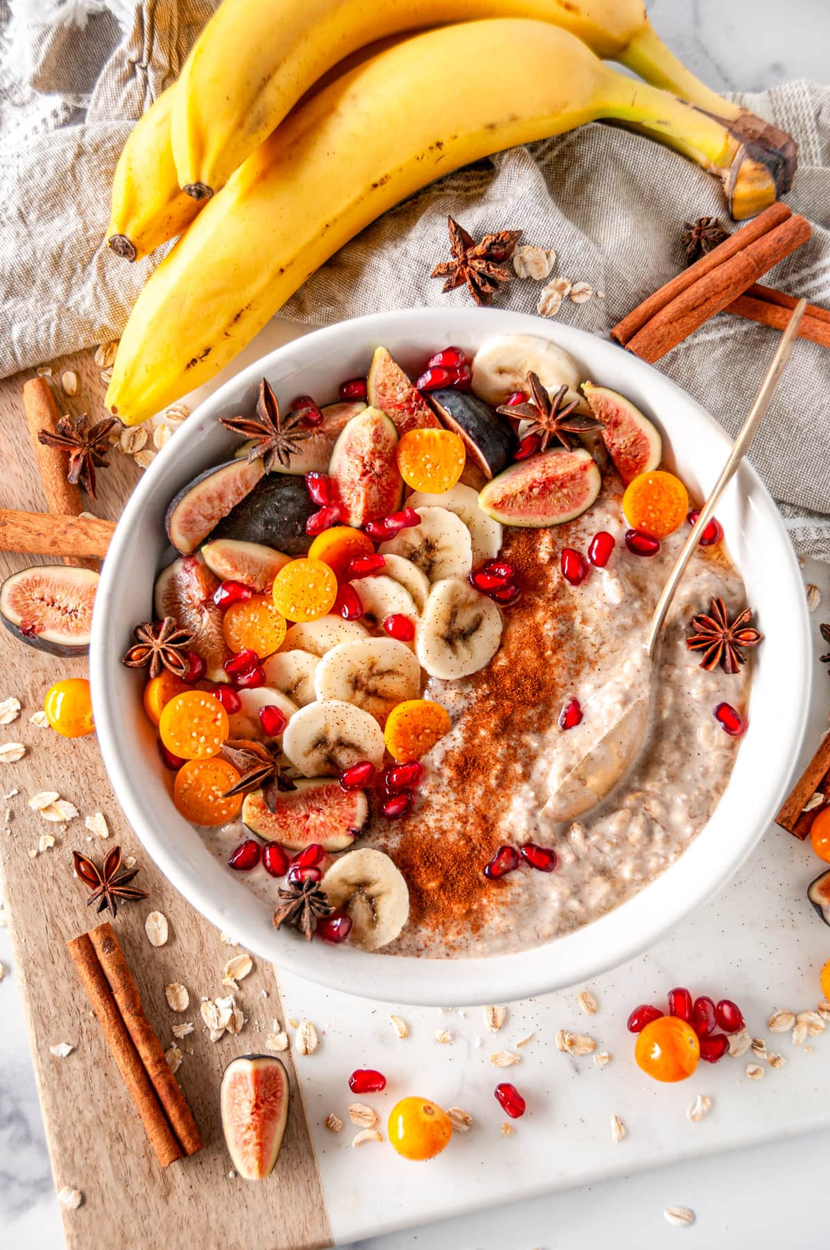 Banana Bread Overnight Oats in white bowl with gold spoon, sliced figs, golden berries, pomegranate seeds and star anise on white marble