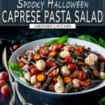Halloween Caprese Pasta Salad in white bowl with silver tongs and cherry tomatoes on a black surface with dark wood window background - white text overlay