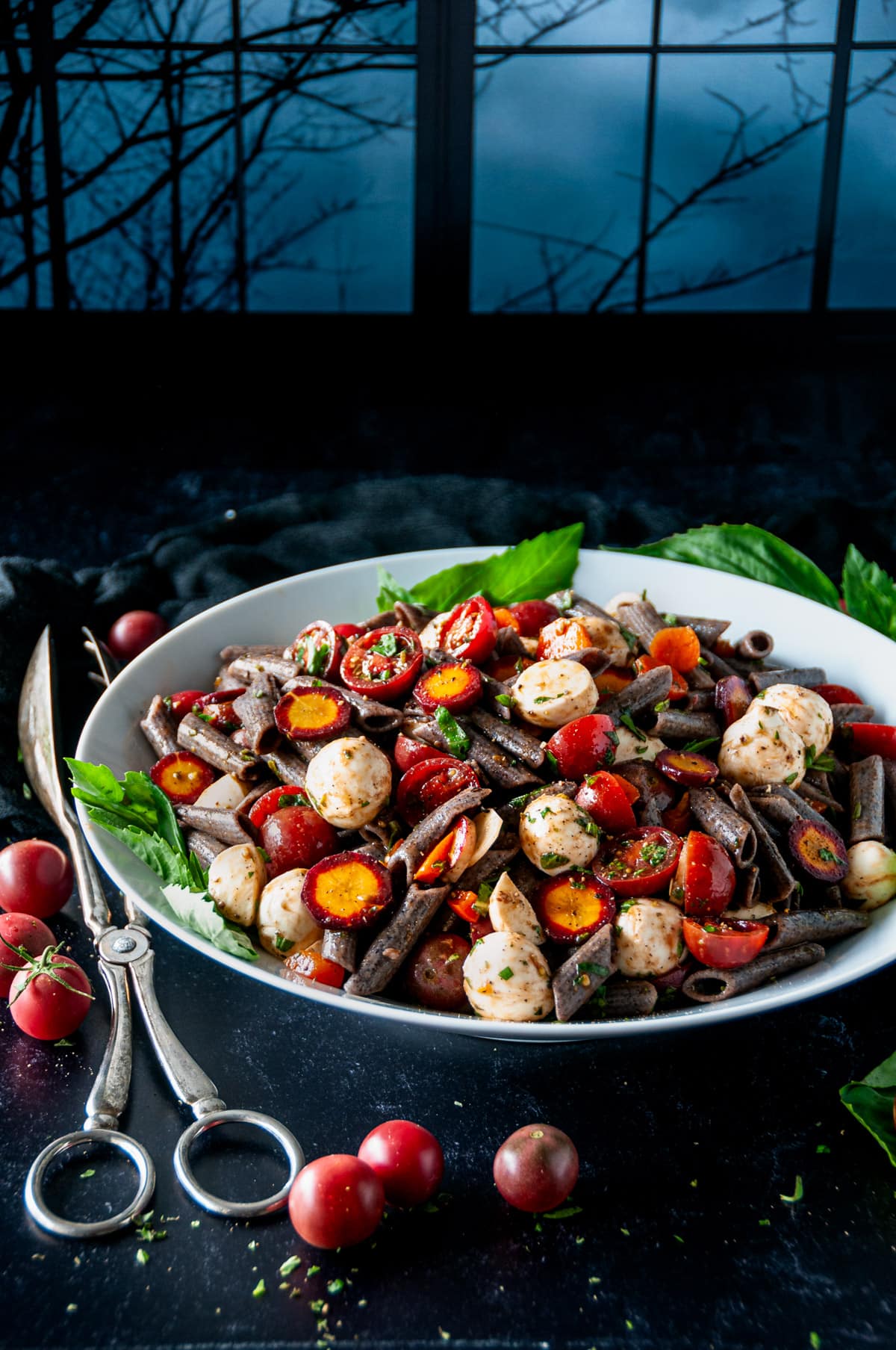 Halloween Caprese Pasta Salad in white bowl with silver tongs  and cherry tomatoes on a black surface with dark wood window background