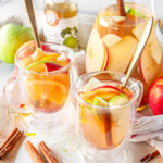 Spiced Apple Cider Sangria in glass mugs and pitcher with orange slices, apple slices, pomegranate seeds and cinnamon sticks on white marble.