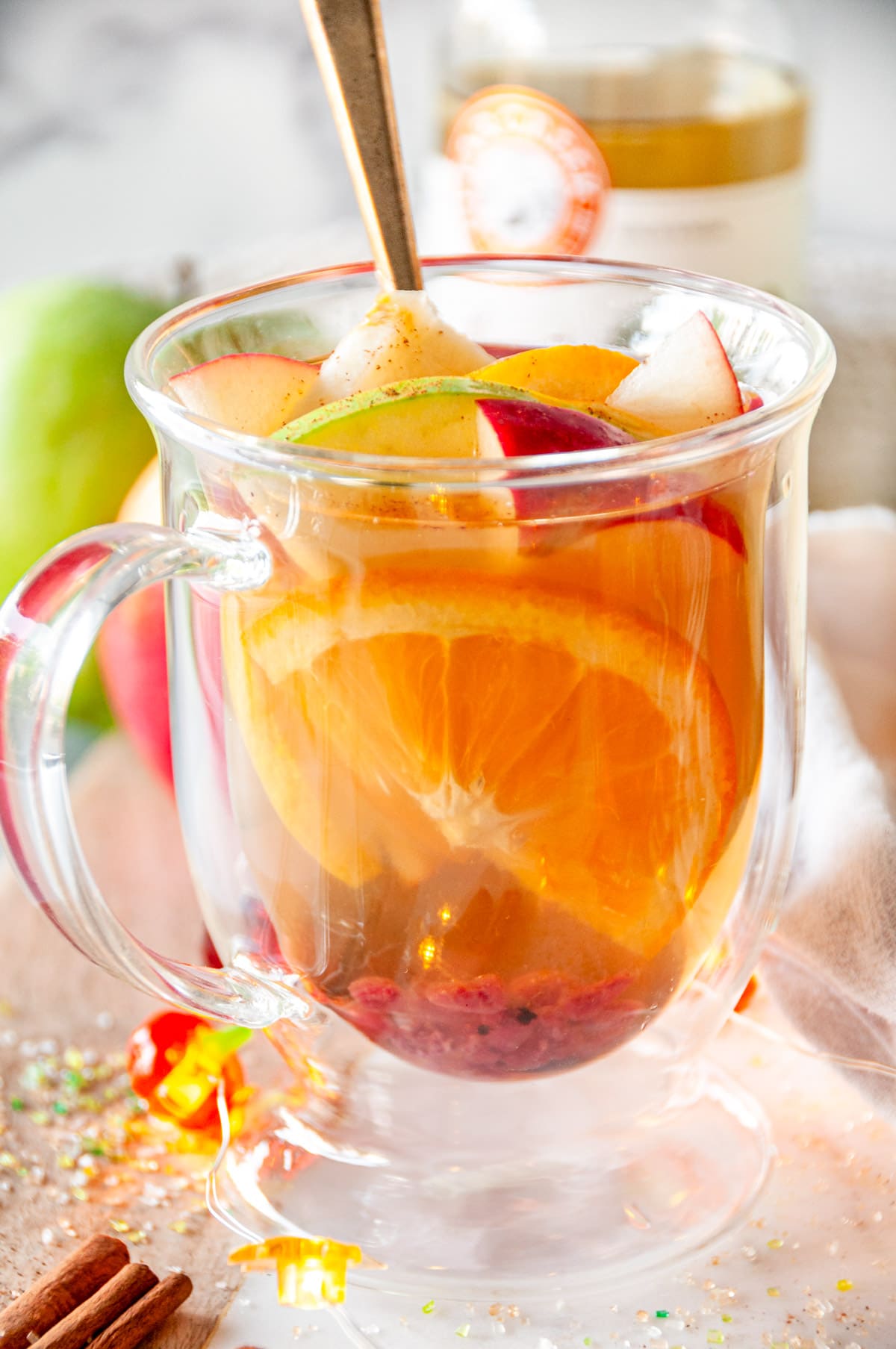 Spiced Apple Cider Sangria in glass mug with orange slices, apple slices, pomegranate seeds and cinnamon sticks on white marble.