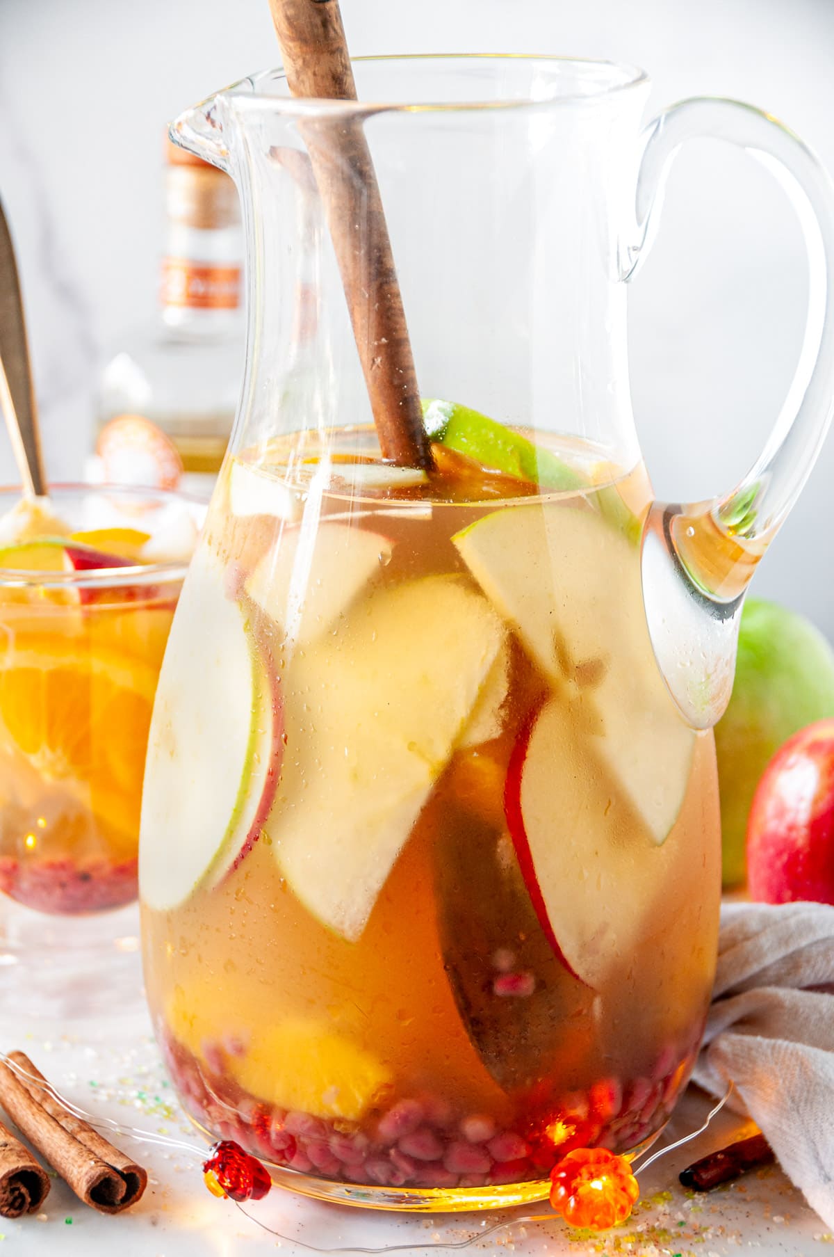 Spiced Apple Cider Sangria in glass pitcher with orange slices, apple slices, pomegranate seeds and cinnamon sticks on white marble.
