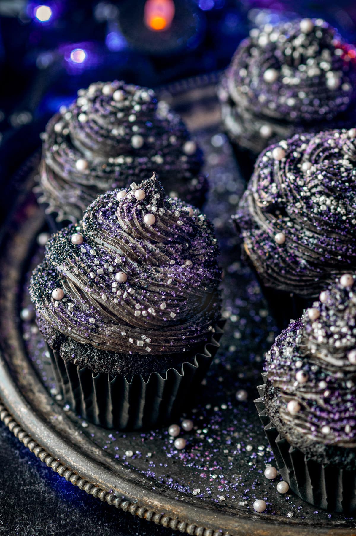 Black velvet cupcakes topped with sanding sugar and sprinkles on gray metal plate with purple lights and candles in background.