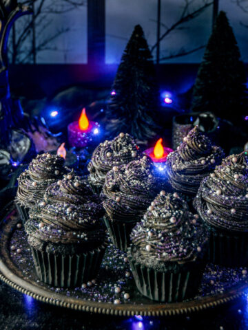 Black velvet cupcakes topped with sanding sugar and sprinkles on gray metal plate with purple lights, candles, and eerie forest window moonscape in background