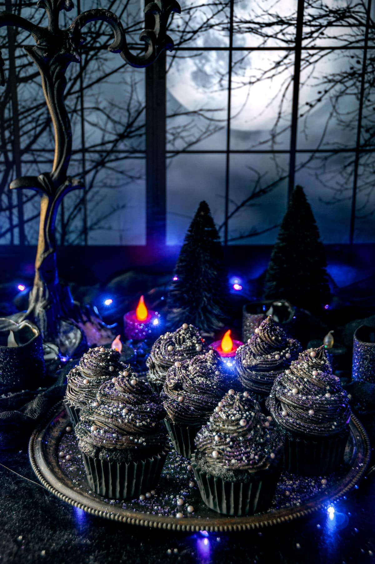 Black velvet cupcakes topped with sanding sugar and sprinkles on gray metal plate with purple lights and candles in background.