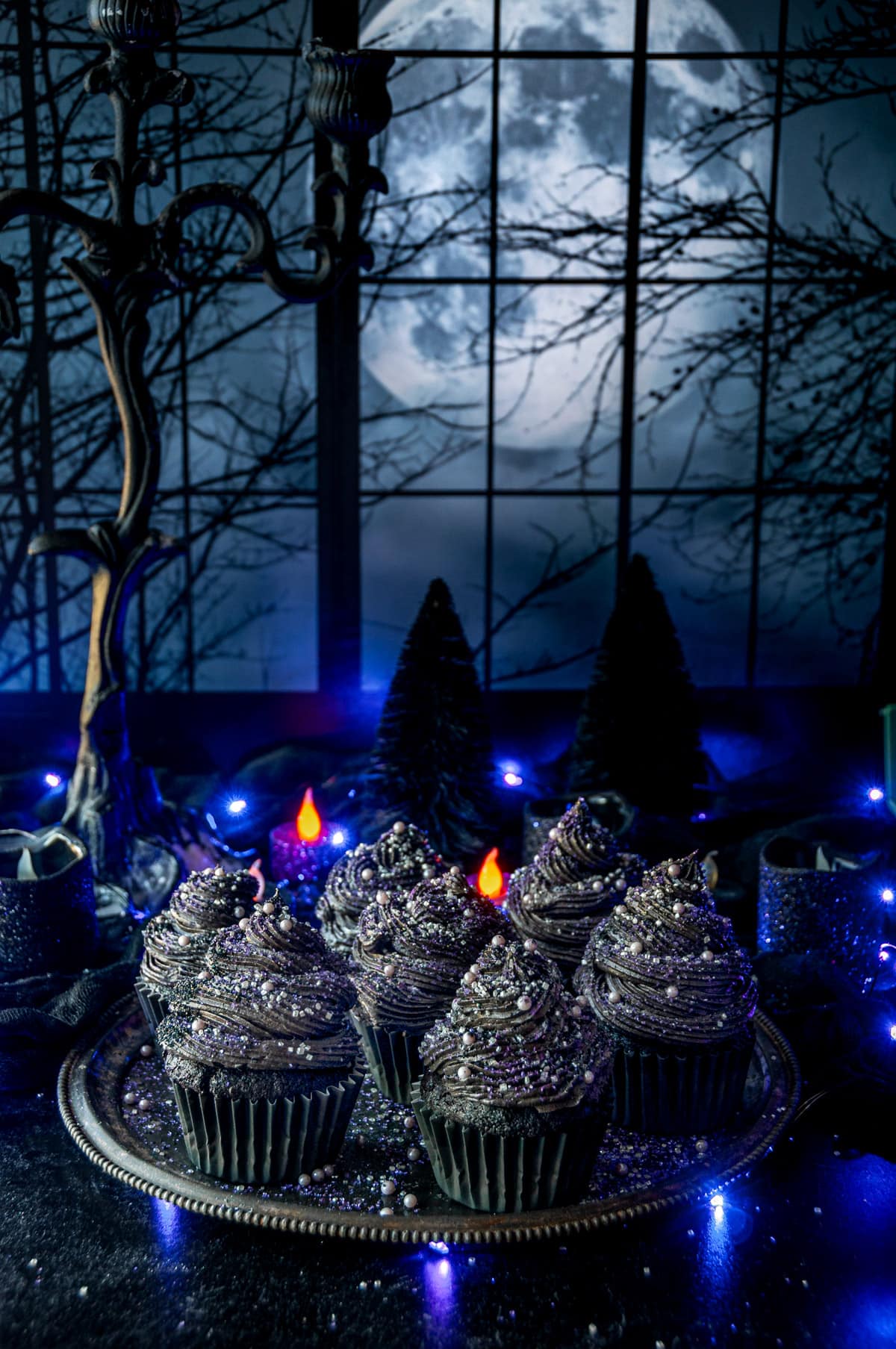 Black velvet cupcakes topped with sanding sugar and sprinkles on gray metal plate with purple lights, candles, and eerie forest window moonscape in background