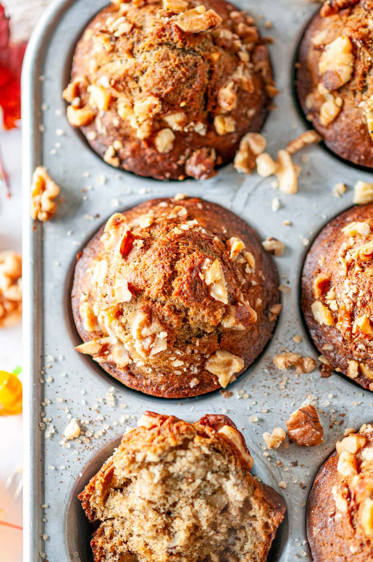 Maple banana muffins in gray metal muffin pan with walnuts and leaves on white marble